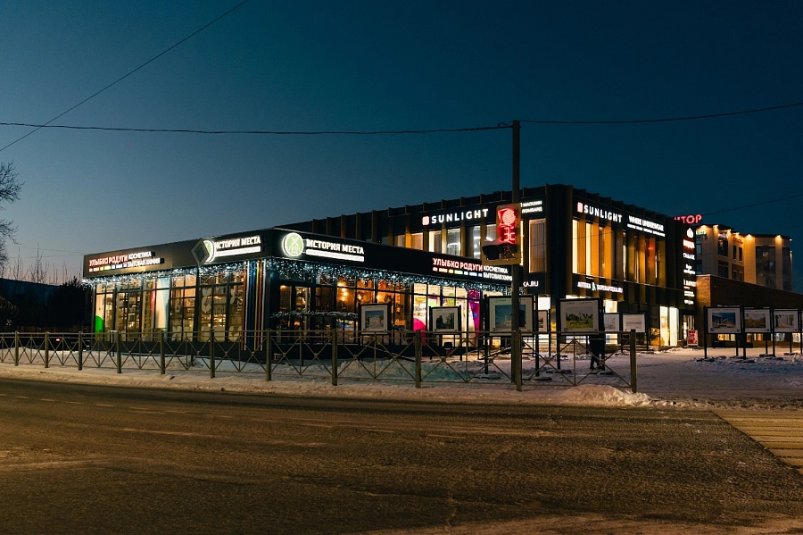 Здание с кофейней «История места» и магазинами на освещённой улице зимой, вечерний городской пейзаж.