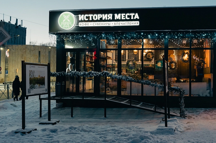 Кофейня и сувенирная лавка «История места» зимой, украшенная огоньками, с панорамными окнами и входом с пандусом.