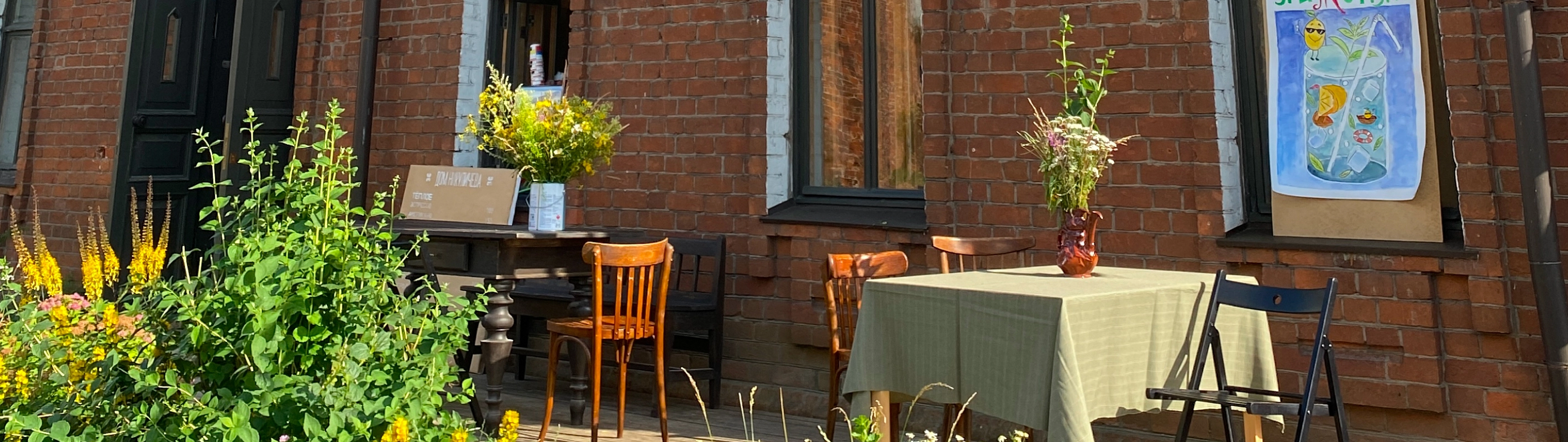 Летняя терраса с деревянными столами и букетами цветов у фасада кирпичного дома.