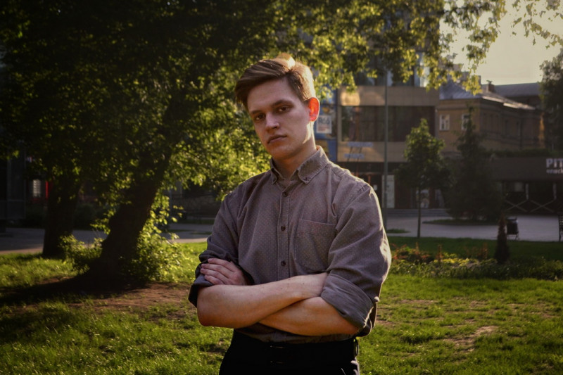 Молодой человек стоит с перекрещенными руками на фоне городской зелени