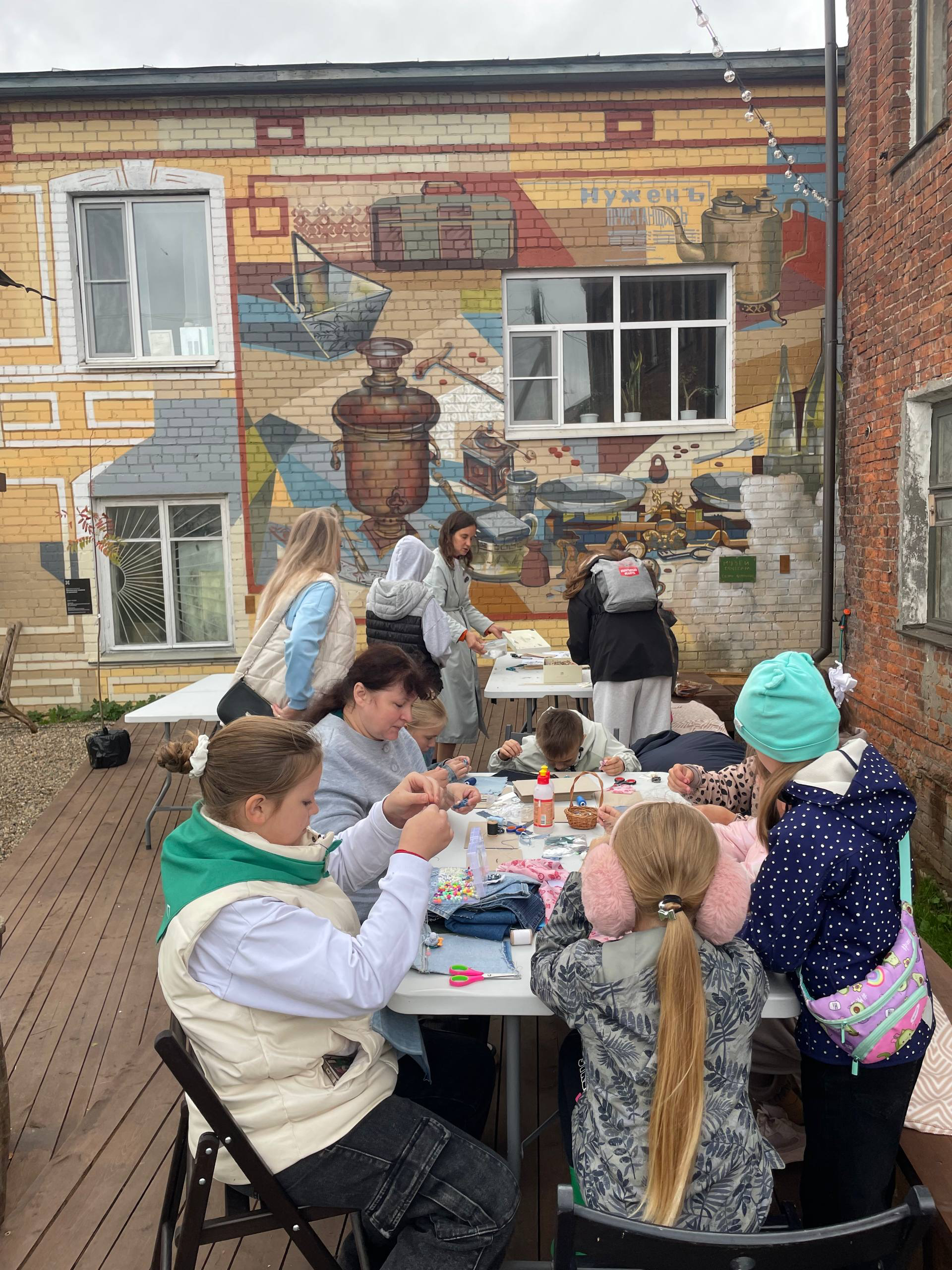 Творческий мастер-класс для детей и взрослых за длинным столом во дворе с ярким настенным муралом.