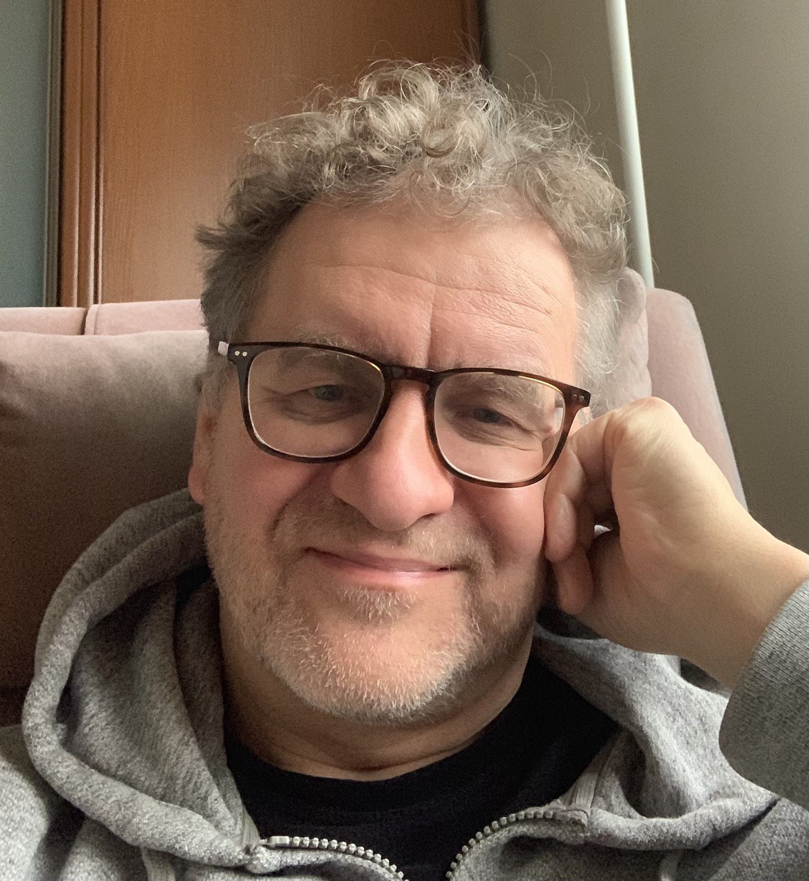 Мужчина в очках и серой толстовке сидит в кресле и улыбается