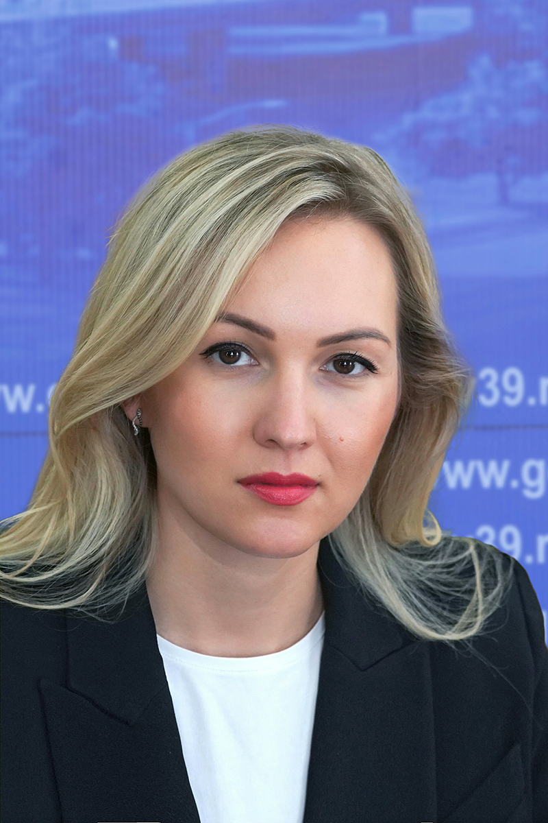 Женщина с блондинистыми волосами в черном пиджаке и белой блузке позирует на фоне синего баннера с текстом.