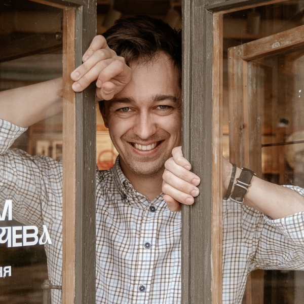 Портрет мужчины в клетчатой рубашке, улыбающегося в дверном проёме.