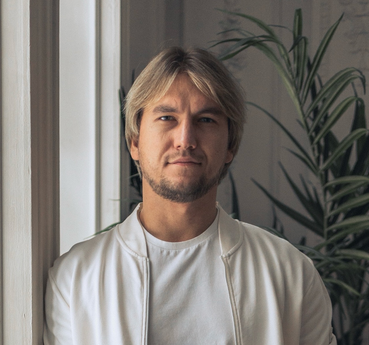 Портрет молодого мужчины со светлыми волосами и бородой в белой футболке и куртке на фоне зелёного растения.