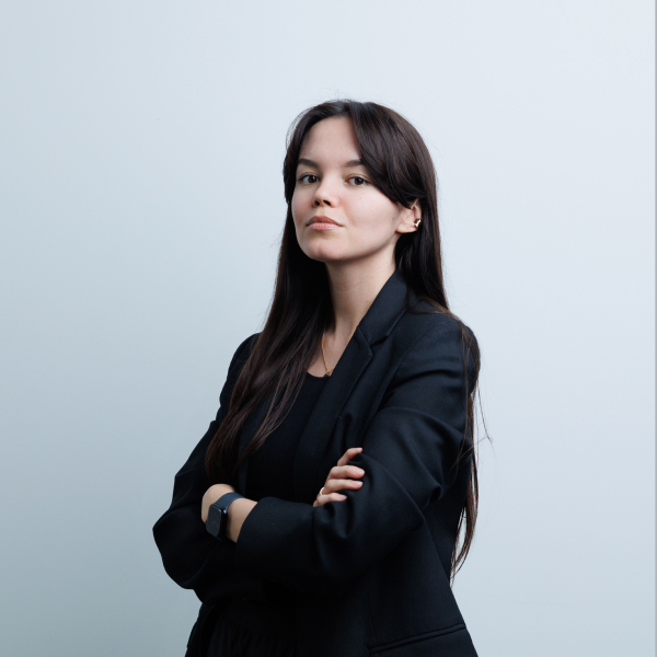 Молодая женщина в чёрном пиджаке позирует на фоне светлой стены, деловой портрет.