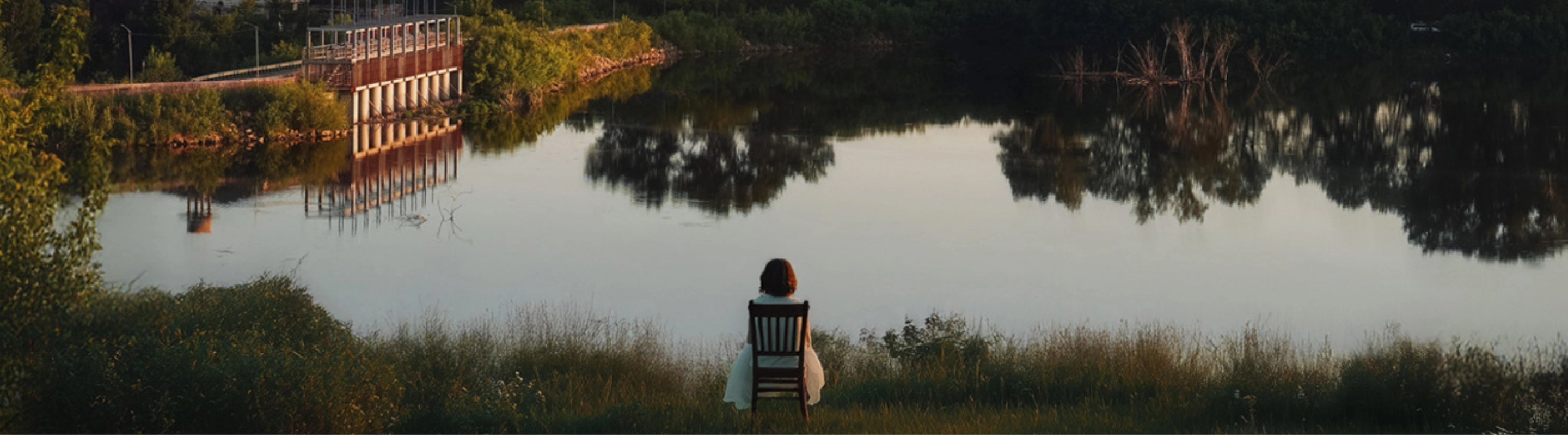 Женщина в белом платье сидит на стуле у воды, перед мостиком и деревьями.