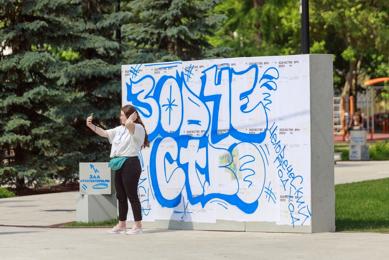 Девушка делает селфи на фоне граффити с надписью Зодчество ВРН в городском парке